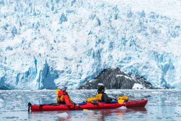 Two people in a red double kayak look at Aialik Glacier on a guided kayak tour