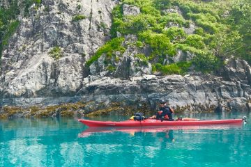 A kayak guide in a single kayak glides through turquoise waters near the coastline
